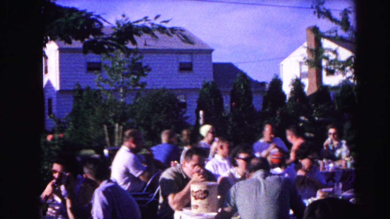 Adults seated at outdoor tables at a backyard cookout, white residential house and evergreen trees in background.
