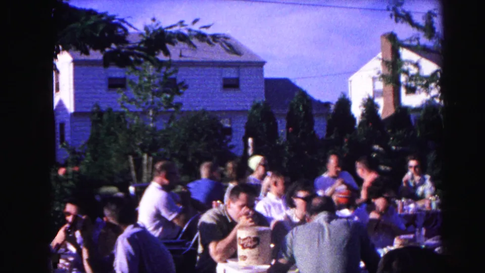 Group of adults gathered at a backyard cookout in Hagerstown, Maryland, circa 1963; white multi-story house and conifers behind them.