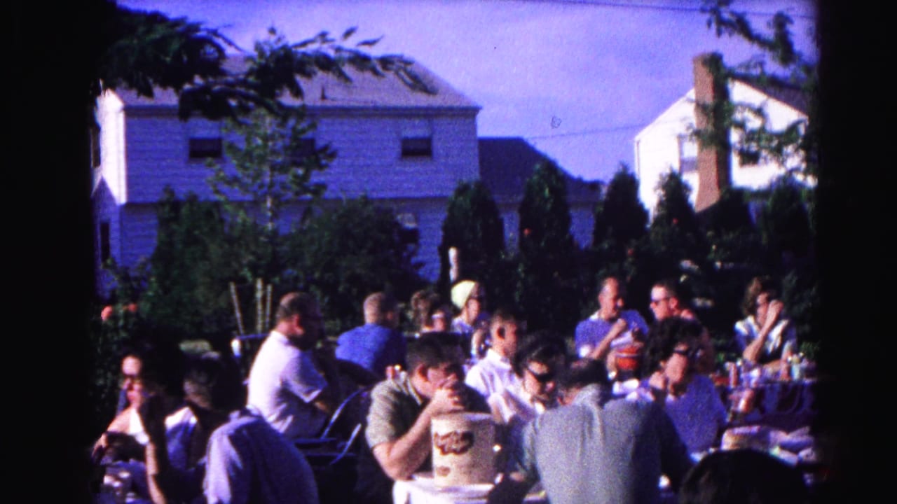Adults eating and socializing at outdoor tables in a residential backyard, large cylindrical container visible in the foreground.