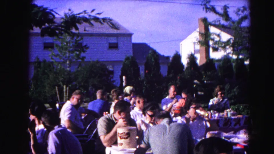 Wide view of backyard cookout gathering, Hagerstown, Maryland, circa 1963; multiple adults at tables with conifers and residential homes behind.