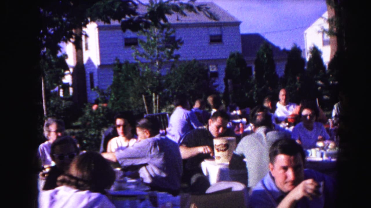 Adults at outdoor tables; man in light blue shirt prominent in foreground right, trees and white suburban building behind.