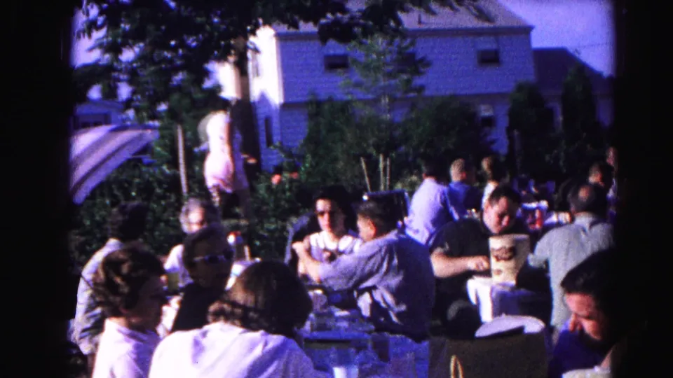 8mm home movie frame, c.1963, closer view of communal outdoor gathering beneath a striped umbrella; adults eating and socializing at tables.