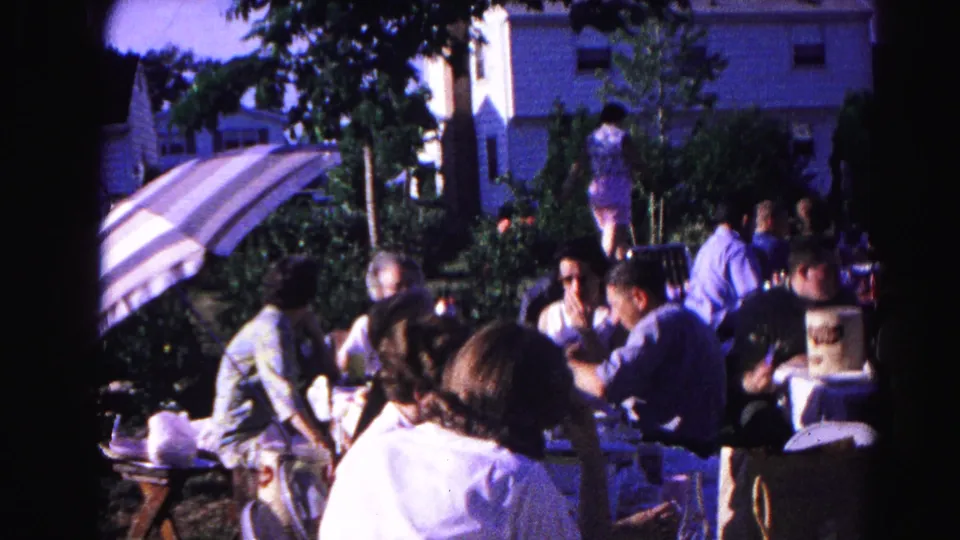 8mm home movie frame, c.1963, woman in white top at the foreground of a communal outdoor gathering; striped umbrella and suburban building behind.