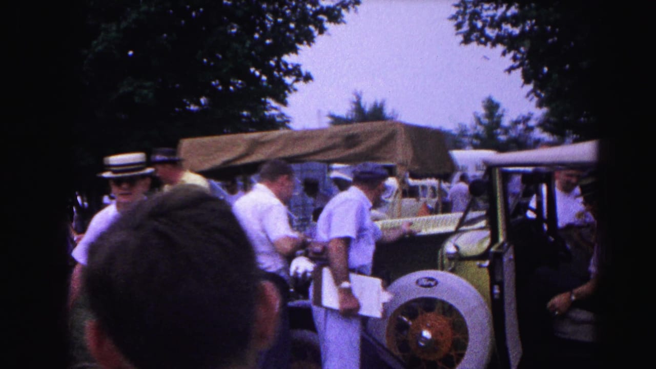 Man in a straw boater hat prominently positioned among a group inspecting a yellow-green vintage open-top automobile, consistent with an early-1960s outdoor car show, Hagerstown, Maryland.