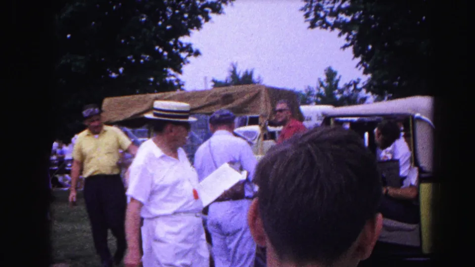 Wider crowd view around a yellow-green open-top vintage automobile with door open, man in yellow shirt at left and boater hat visible at center, with background figures, circa 1963 outdoor community event.