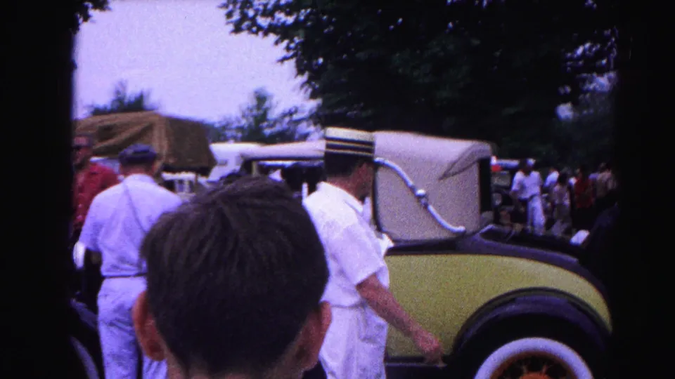 Handheld over-shoulder shot past an unidentified foreground figure toward a man in a straw boater hat beside a yellow-green vintage automobile, with a larger crowd and trees visible at right, circa 1963.