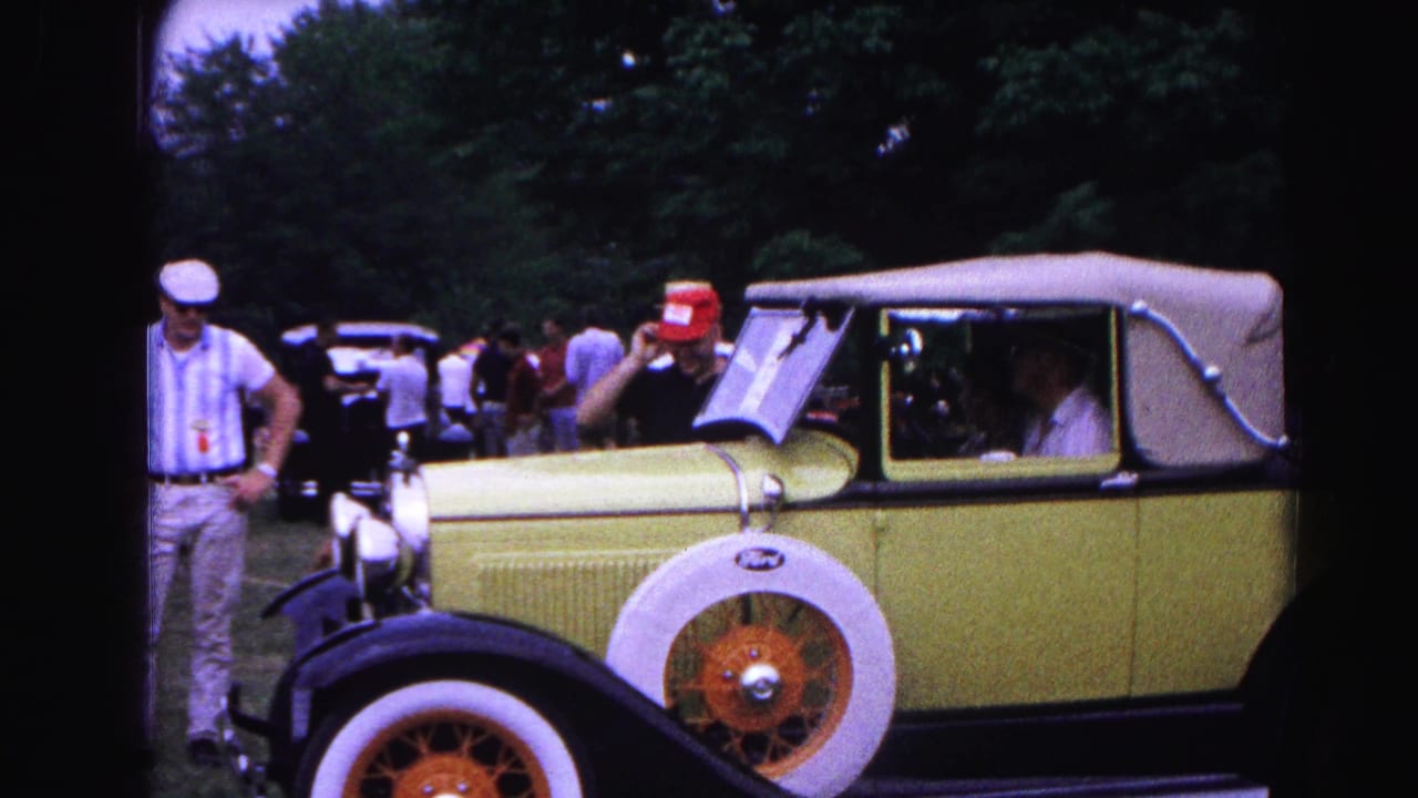 Side view of a yellow Ford convertible with a man in a red cap beside it.