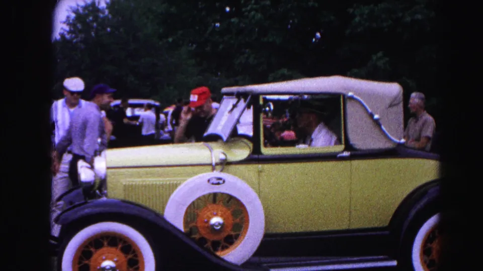 The yellow Ford convertible is seen in profile on the grass, with visitors clustered near the front and a seated driver or passenger visible through the side window.