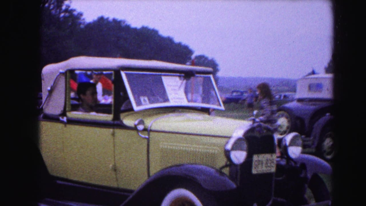 Pale yellow pre-war convertible with white soft top and a windshield display card at a 1963 classic car show in Hagerstown, Maryland; attendees and black vintage cars in background.