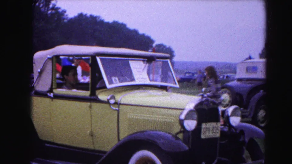 Pale yellow pre-war convertible with white soft top and a windshield display card at a 1963 classic car show in Hagerstown, Maryland; attendees and black vintage cars in background.