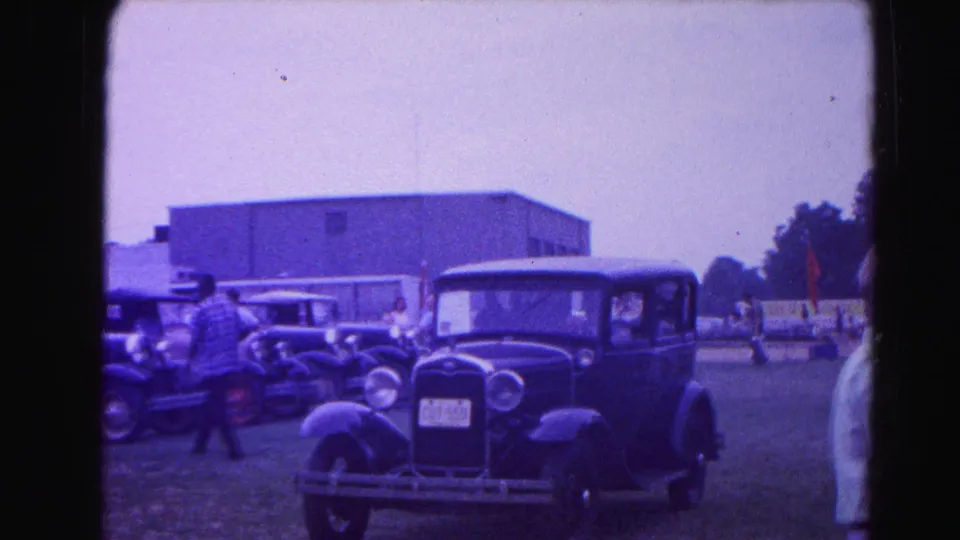 Wide field view of the 1963 Hagerstown, Maryland classic car show: pre-war touring cars lined on grass with 1960s attendees browsing; aged 8mm stock gives a pronounced cool-blue cast.