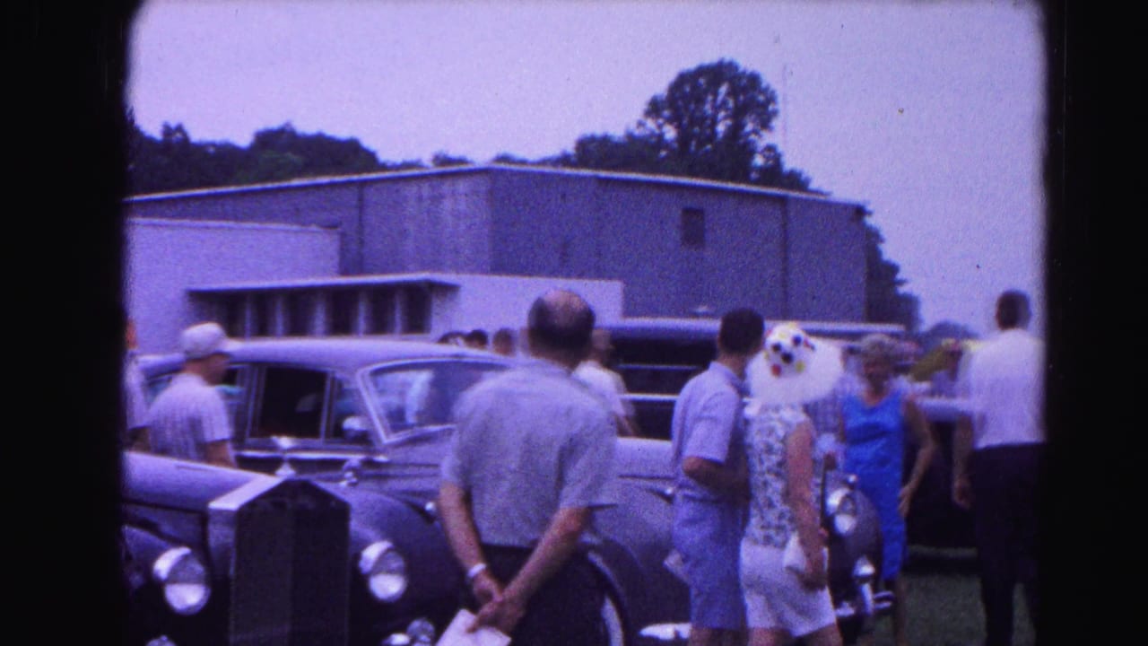 Group of adults in summer clothing standing near large dark classic cars on a grassy lot, flat-roofed building behind them.