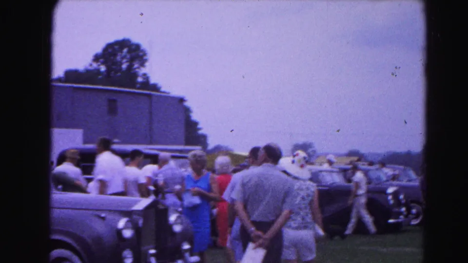 Wide shot of a grassy outdoor car show with groups of adults in early-1960s attire strolling past a row of classic vehicles; consistent with 1963 Hagerstown, Maryland.