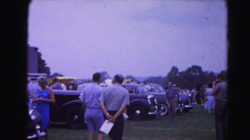 Wide view of a car show crowd walking beside a long row of classic automobiles on a grassy field; 8mm footage consistent with 1963 Hagerstown, Maryland.