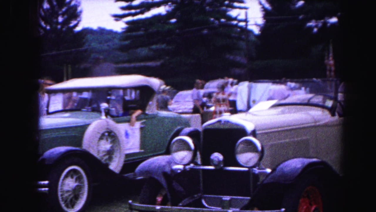 Close view of a cream antique roadster with round headlights and red wire wheels beside a dark green vintage sedan at an outdoor car show.