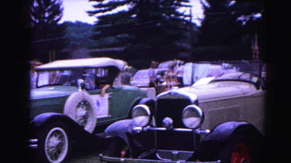 8mm frame from a 1963 Hagerstown, Maryland car show: cream open-top antique roadster and green sedan on grass, spectators and conifers in background.