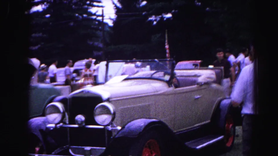 8mm home-movie frame, 1963 car show: man examining the hood of a cream antique convertible, spectators and a small flag visible in background.