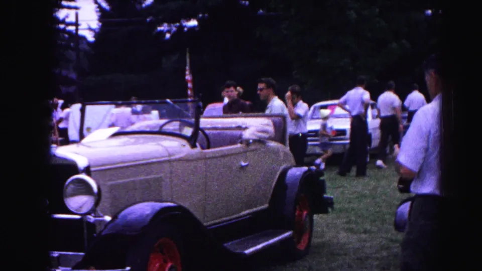 8mm frame from 1963 Hagerstown car show: side profile of cream antique roadster with red wheels, adults in summer dress walking the show grounds.