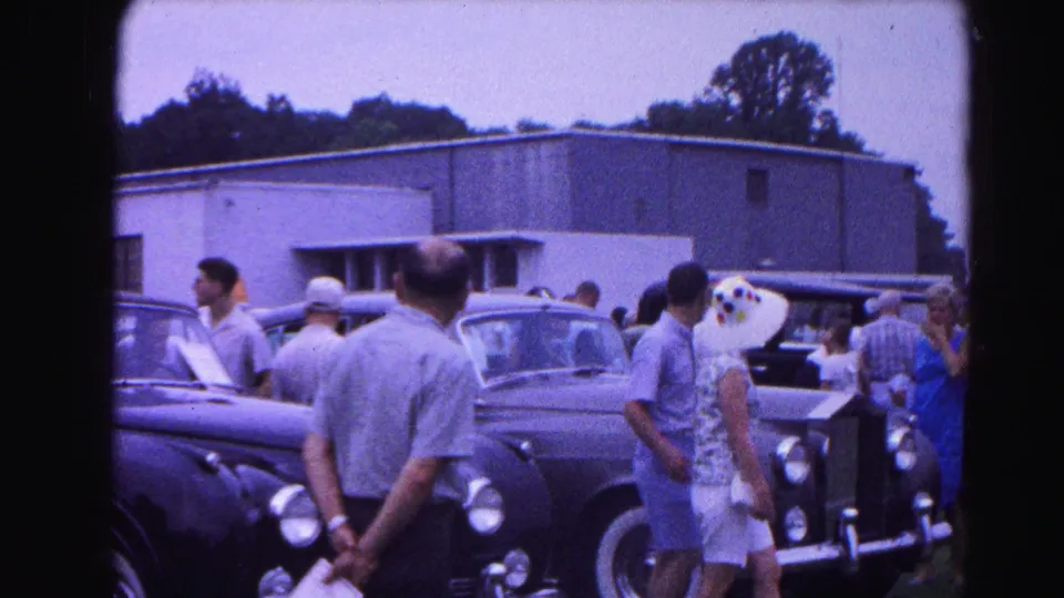 8mm frame from 1963 Hagerstown car show: spectators including a woman in a blue dress and a man in a grey shirt examine vintage automobiles, flat-roofed building behind.