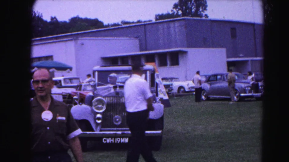 1963 Hagerstown, Maryland car show: attendee in dark shirt beside a light blue Rolls-Royce on a grassy field; other 1960s visitors and vehicles behind.