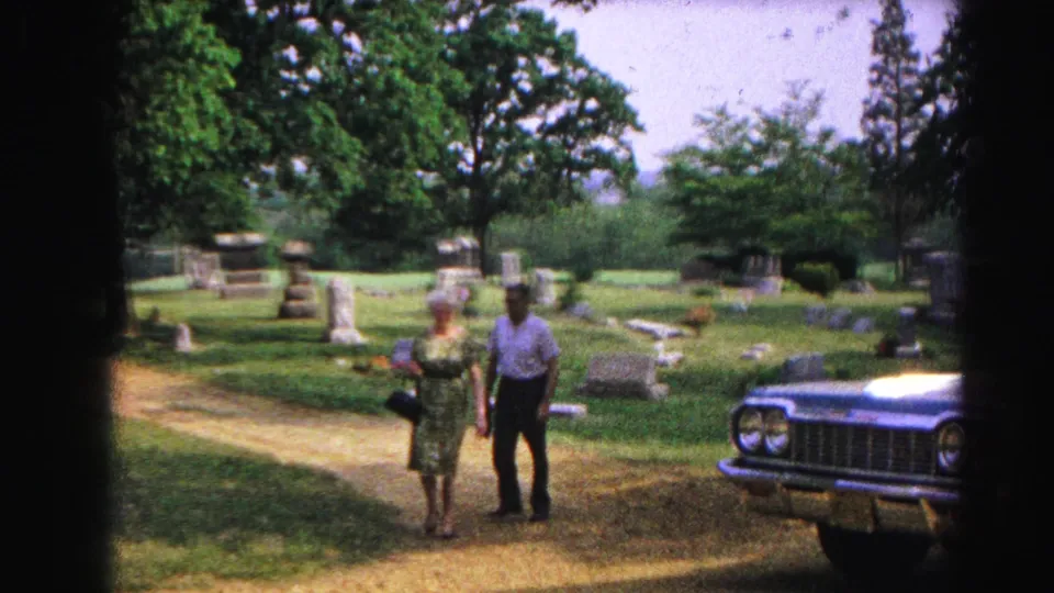 Man and woman in early-1960s clothing walking through a cemetery setting circa 1963; large dark American sedan prominent in foreground, gravestones and green trees behind.