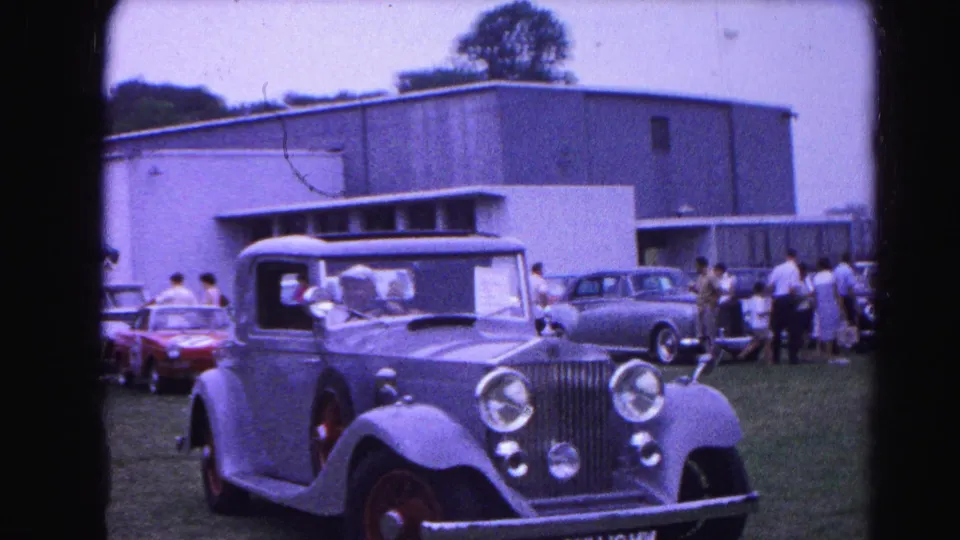 Outdoor classic car show circa 1963, possibly Hagerstown Maryland; a pre-war open-top luxury automobile on grass with visitors browsing and a flat-roofed mid-century building in background.