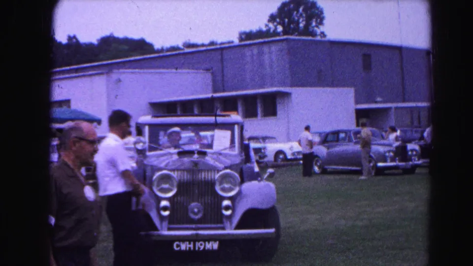 Attendee examining a pre-war luxury automobile at an outdoor classic car exhibition circa 1963; vehicle registration plate visible on car; mid-century building and other classic cars in background.