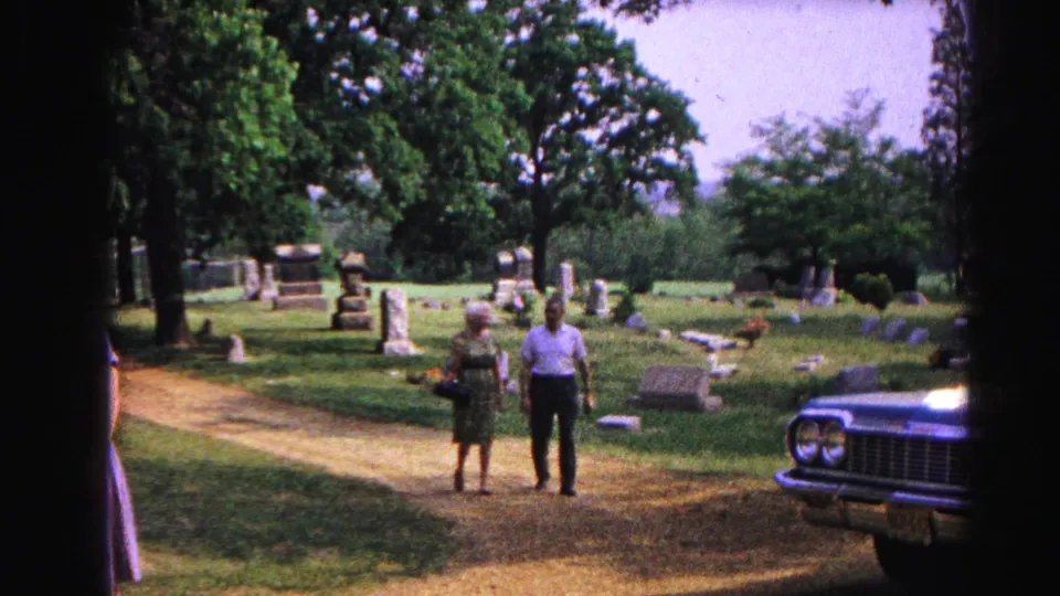 Multiple adults in period dress walking through a sunlit cemetery toward the camera; a large 1960s-era American car is partially visible at right.