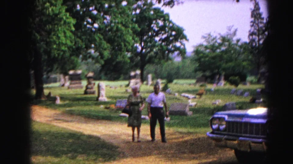 Two women in 1960s-era dresses walk a cemetery dirt path toward the camera; a large dark American sedan is parked at right.