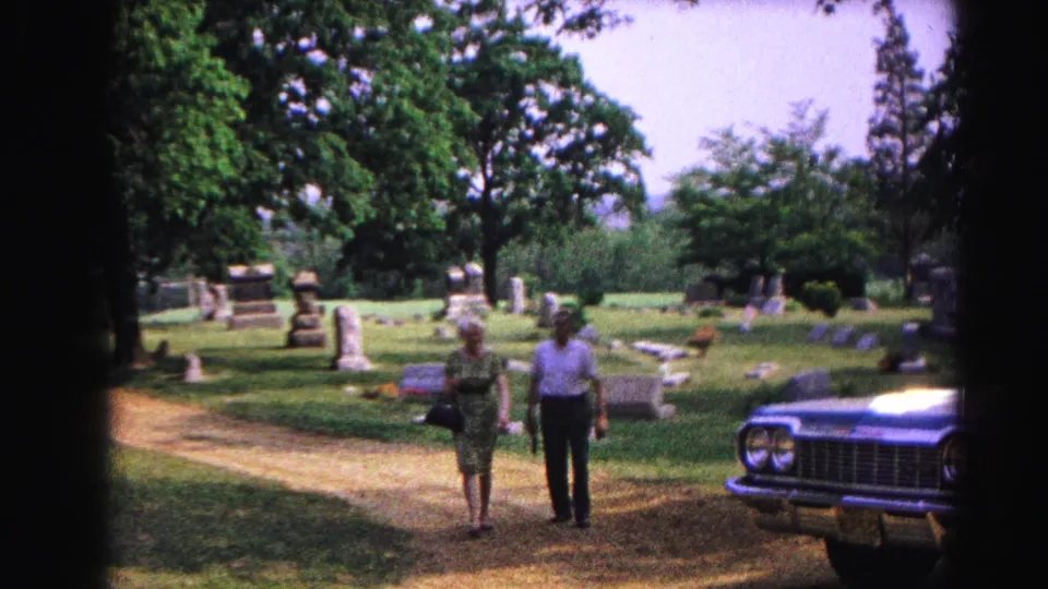 Two adults near the front of a large dark early-1960s American automobile in a cemetery; upright gravestones and mature trees fill the background.