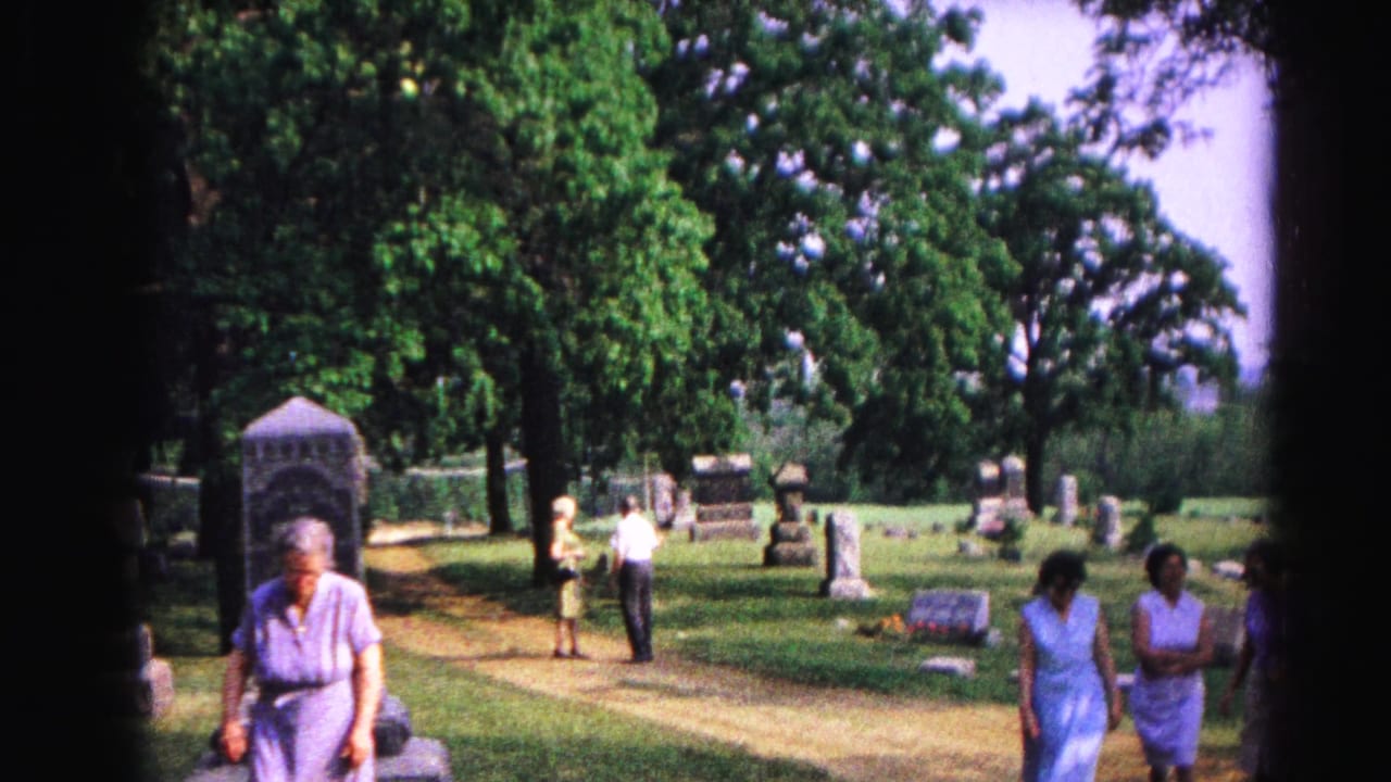 Elderly woman in lavender dress walks toward camera in a sunlit cemetery; other figures and headstones are visible behind her.