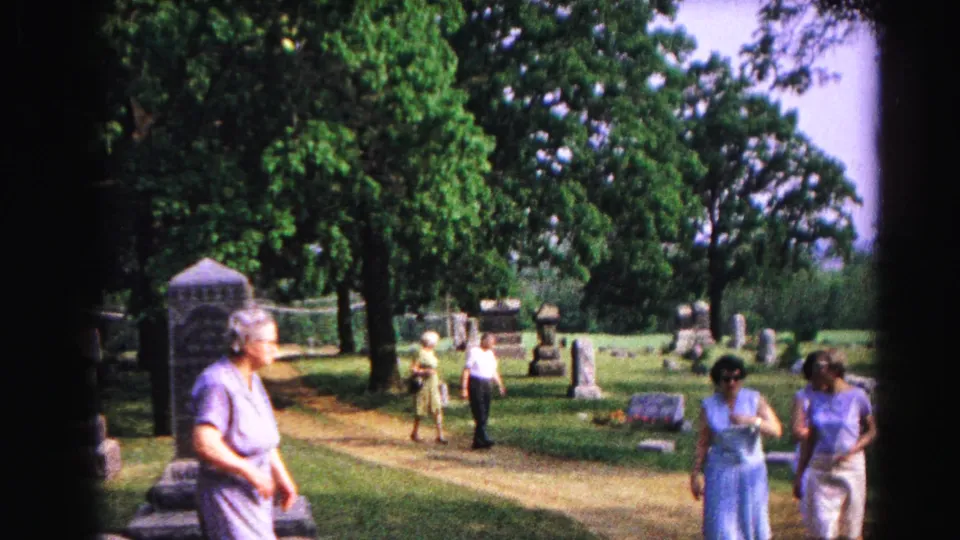 1963 home movie: elderly woman approaches camera in cemetery; two women and a possible child figure visible in the background among headstones and trees.