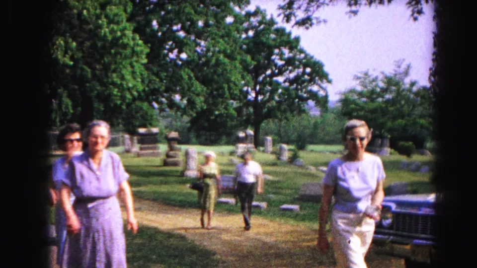 1963 8mm frame: woman in white top moves toward camera alongside others; gravestones, trees, and the partial edge of a dark vehicle visible at right, Hagerstown cemetery.