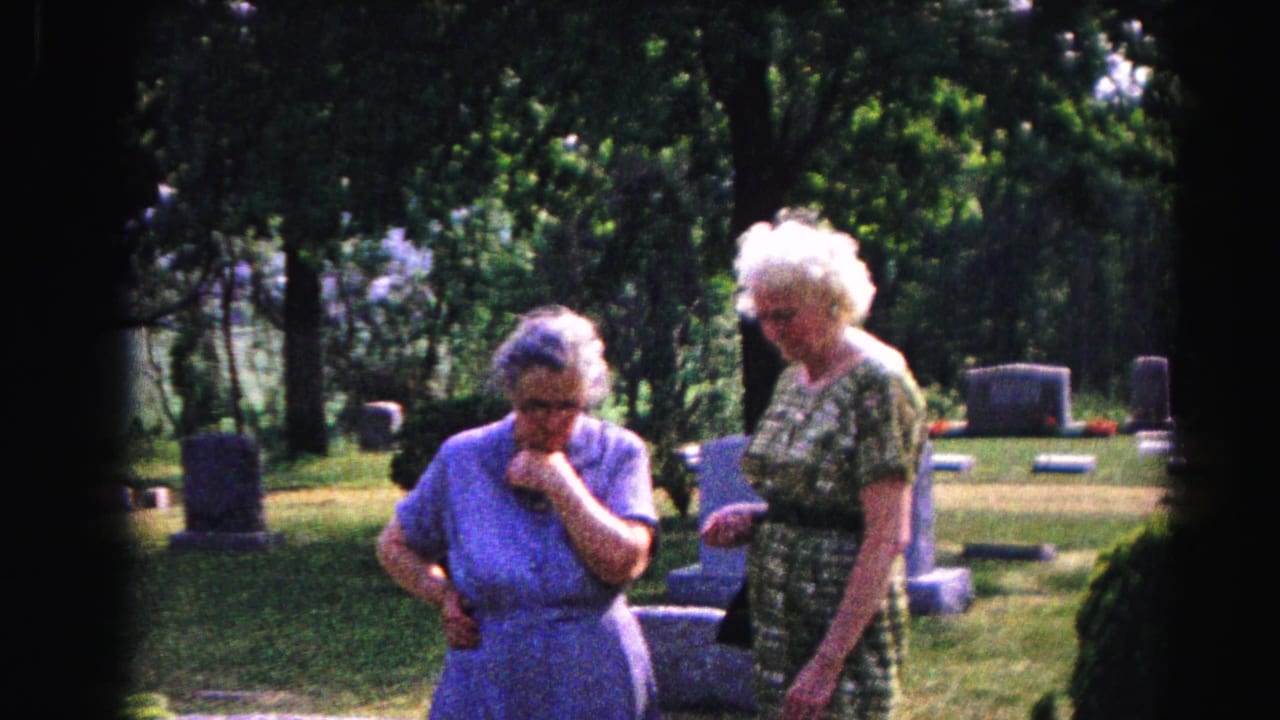 Two elderly women in a cemetery, the woman in blue with hand still near her face, her companion in green patterned dress beside her among headstones.