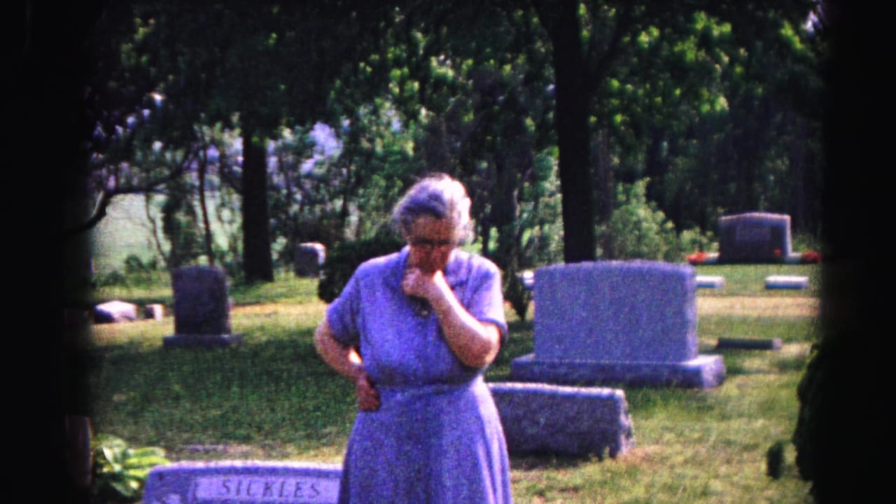 Elderly woman in blue dress stands in cemetery with hand raised to mouth, gravestones and trees behind her.