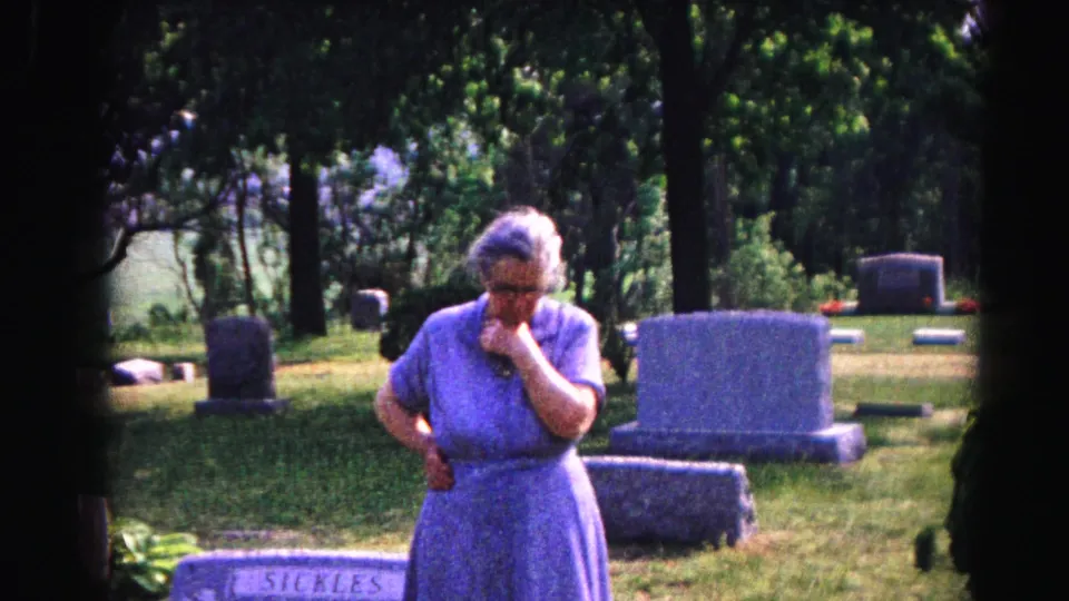 Elderly woman in blue dress holds hand to mouth in grief at a sun-dappled cemetery, circa 1963, Hagerstown, Maryland.