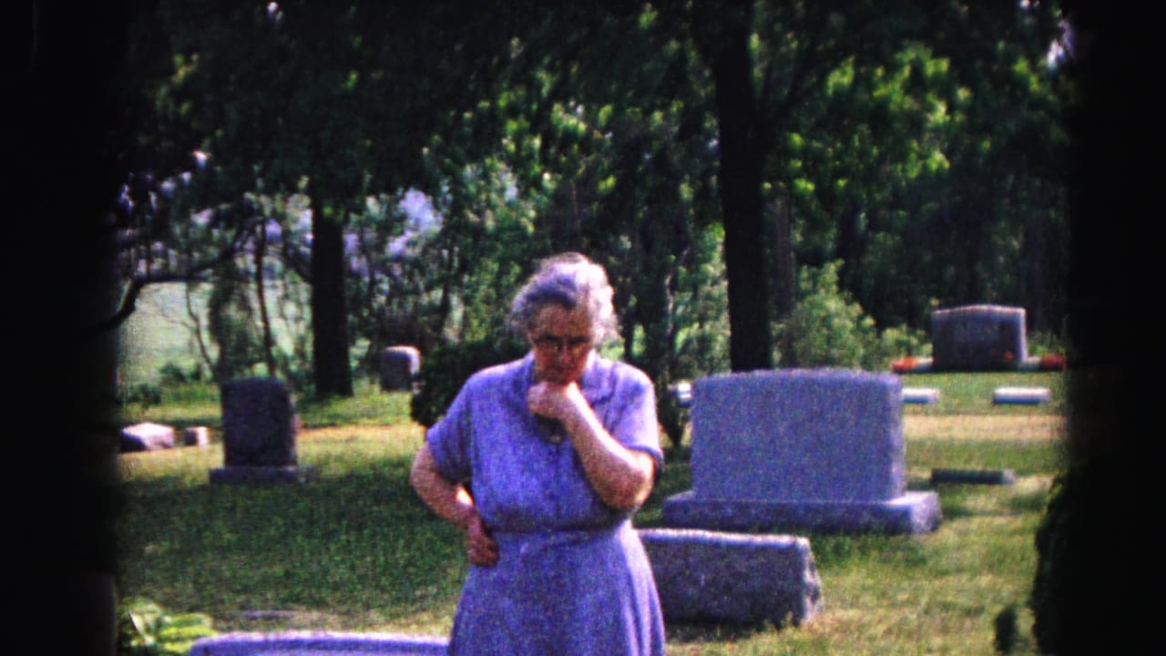 Elderly woman in blue dress at cemetery, hand to mouth, several gravestones visible behind her.