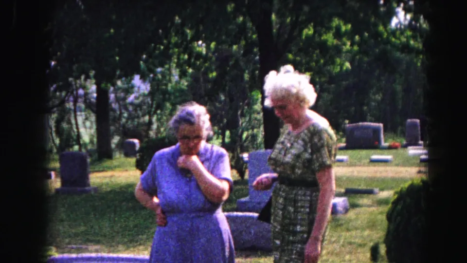 Two elderly women standing together among gravestones in a shared moment of remembrance, 8mm home movie circa 1963, Hagerstown, Maryland.