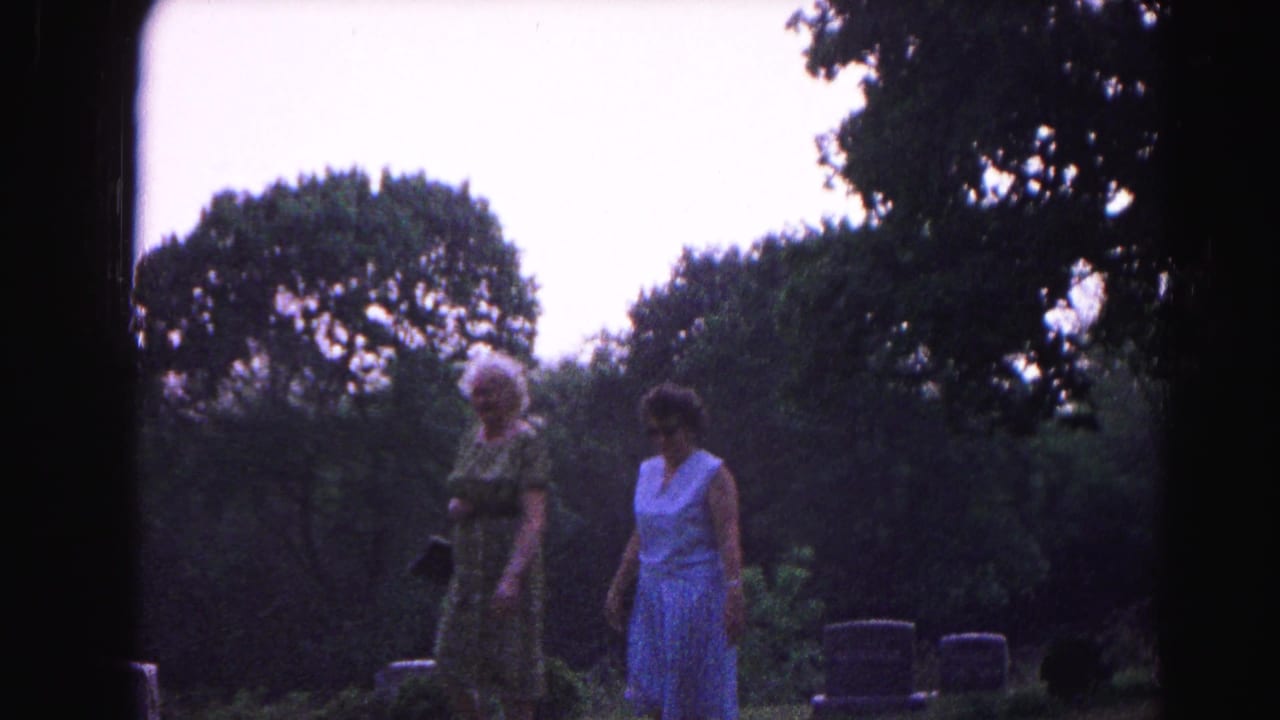 Two women in summer dresses walk among gravestones in a tree-lined cemetery under a pale sky.