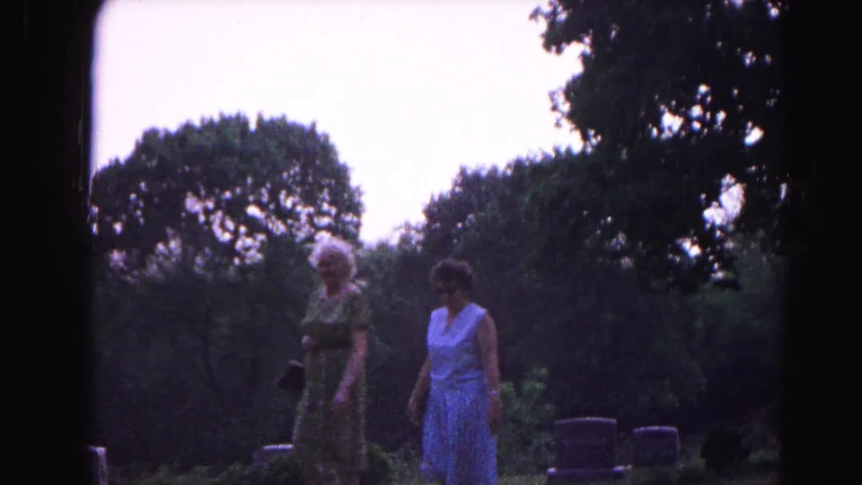 Two women in early-1960s summer dresses walk among upright gravestones in a cemetery in Hagerstown, Maryland, circa 1963, lush deciduous trees behind them.