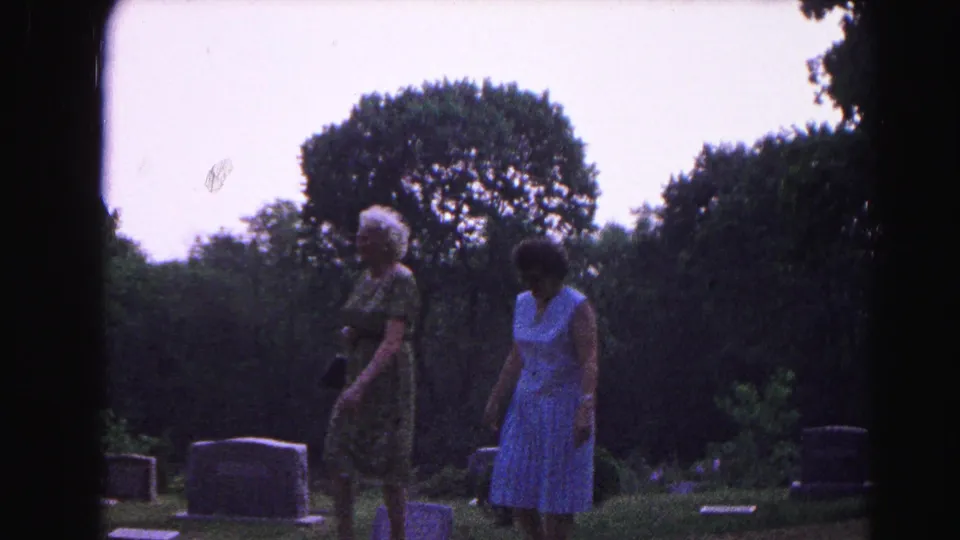 Two women in 1960s summer dresses pause among gravestones in Hagerstown, Maryland, circa 1963, with dense summer trees framing the scene.