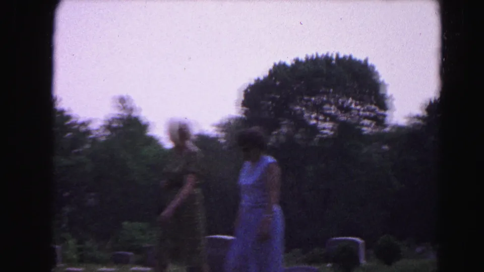 Two women in 1960s dress walk through a cemetery, circa 1963, with a wide overcast sky, scattered gravestones, and motion blur on the closer figure indicating camera movement.