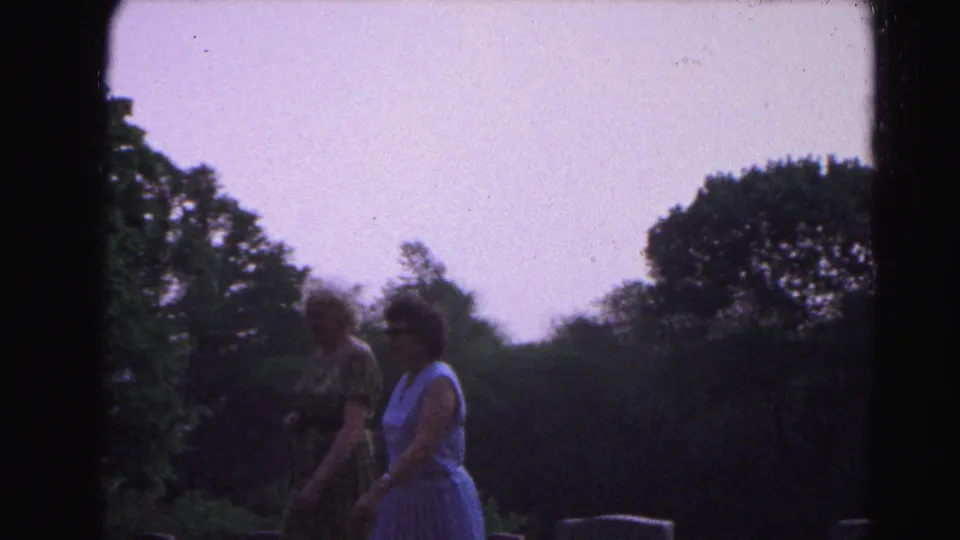 Two women in 1960s summer dresses walk together past gravestones in Hagerstown, Maryland, circa 1963, with summer trees and overcast sky in the background.
