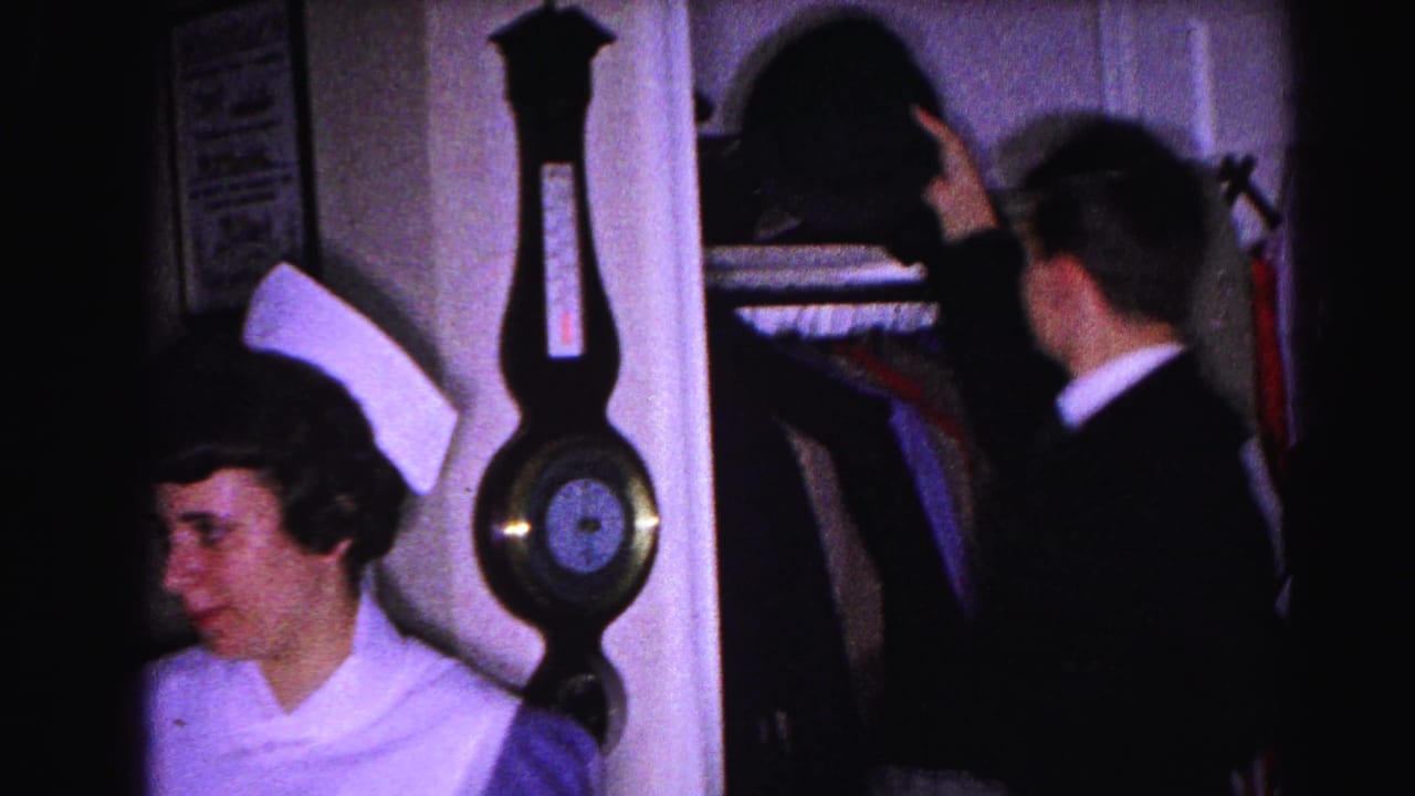 Nurse and man in dark suit standing near an open closet in a home hallway; decorative wall barometer between them.