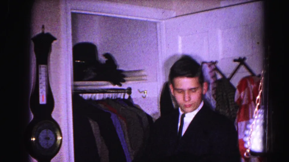 Man in dark suit and nurse in a 1960s home interior standing before an open closet with hanging garments; barometer visible on left wall.