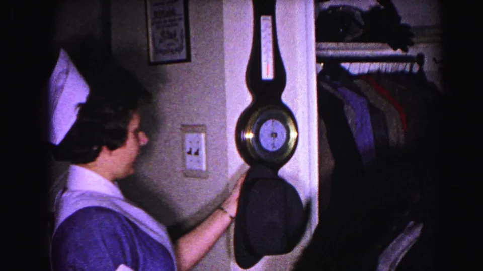 Woman in white domestic uniform viewed from behind, next to a wall-mounted barometer; open closet with hanging coats visible; 1963 home interior, Hagerstown, Maryland.
