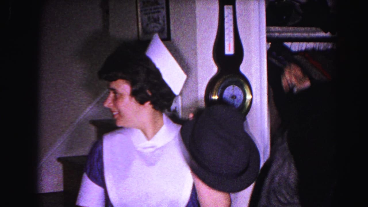 Smiling woman in white maid's uniform and white cap, holding a dark hat, standing in front of a wall barometer and staircase.