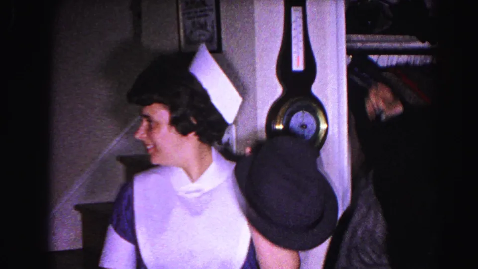 Woman in white maid's uniform and cap smiles toward camera while holding a dark fedora; wall barometer and staircase newel post visible behind her; 1963 Maryland home interior.