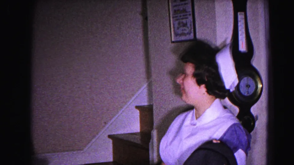 Woman in white domestic uniform in profile beside a staircase, with a large wall-mounted barometer dominating the foreground; framed picture on wall; 1963 home interior.