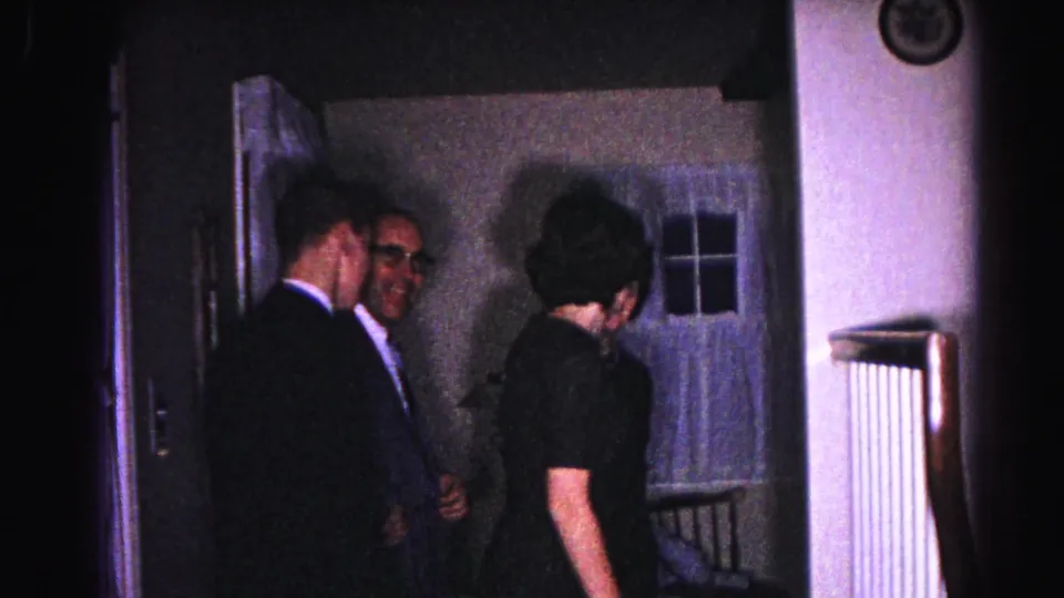 Two men in dark business suits near a residential entry doorway, with a curtained window and wall radiator visible; 1963 home hallway, Hagerstown, Maryland.